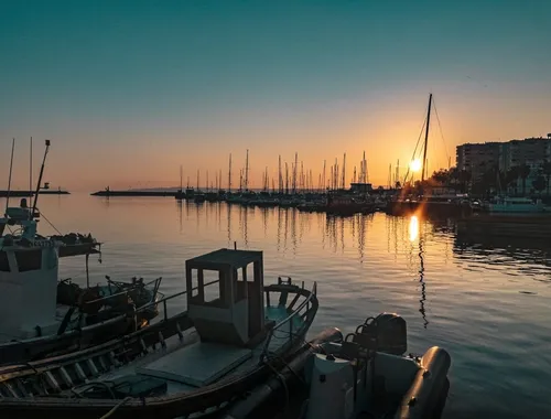 El sol poniéndose detrás de los mástiles de los barcos atracados, pintando el puerto con tonos dorados y naranjas.