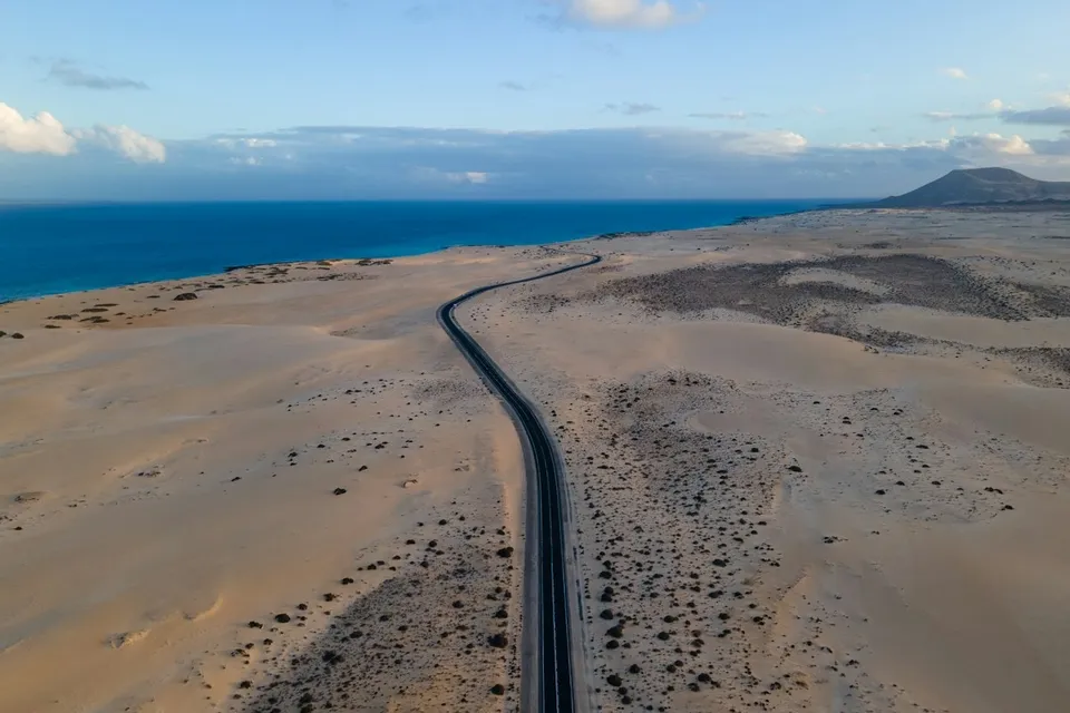 A road crossing the golden dunes next to the blue Atlantic, Fuerteventura. Photo via Unsplash